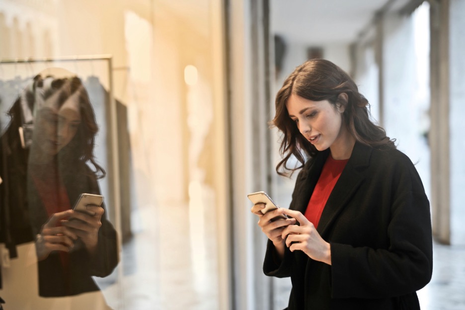 Person standing indoors beside a glass storefront, using a smartphone, with clothing displays and reflections visible nearby.