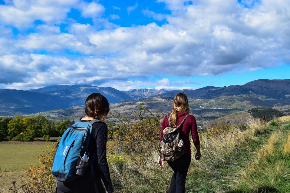 Two people hiking along a grassy trail, wearing backpacks and looking out over rolling hills and distant mountains.
