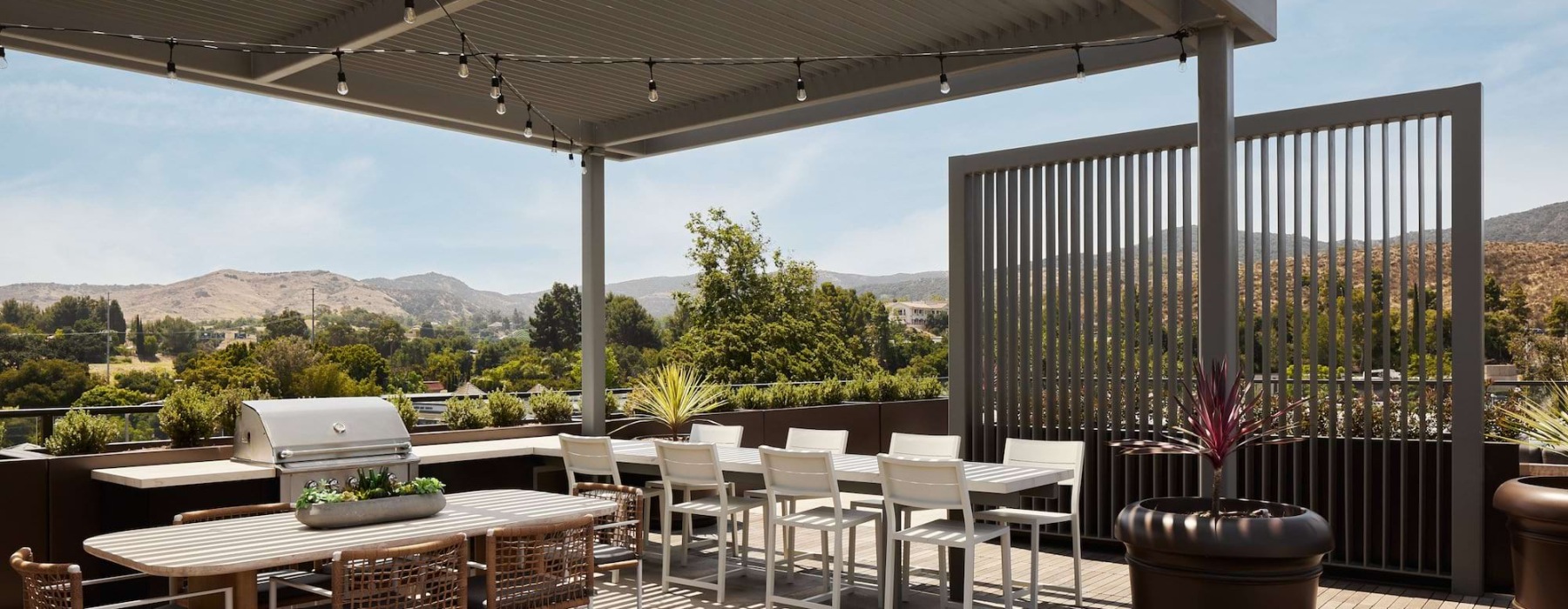 A sunny, modern rooftop patio at Santal Thousand Oaks with a covered dining area, BBQ grill, and views of rolling, tree-covered hills.