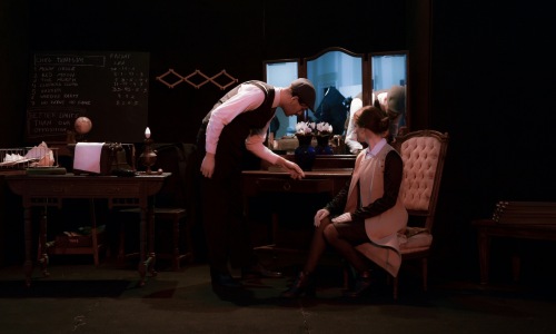 A man wearing a vest and cap leaning toward a woman sitting in a tufted chair on a dark stage near a chalkboard.
