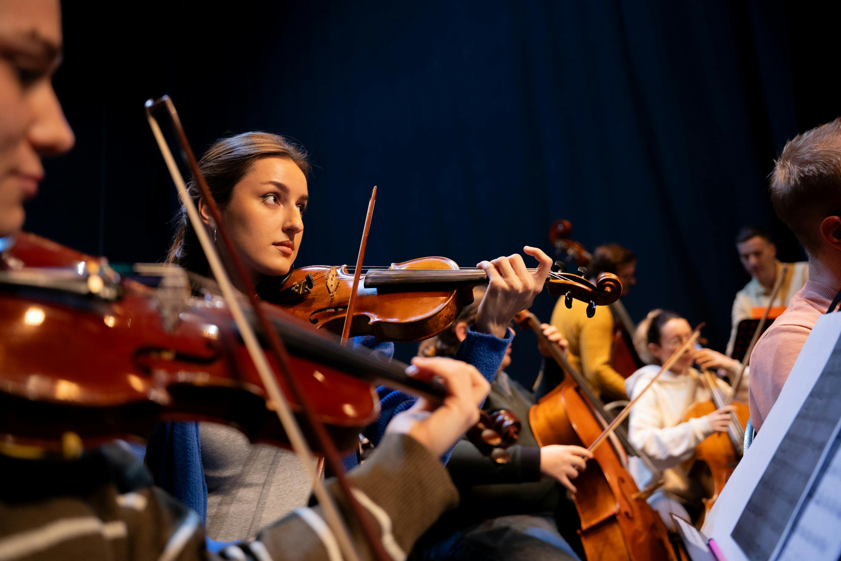 A woman playing a wooden violin alongside other musicians holding cellos and string instruments on a dark stage. A woman playing a wooden violin alongside other musicians holding cellos and string instruments on a dark stage.