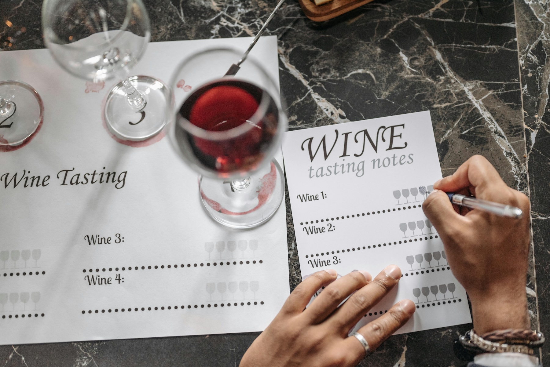 A person holding a pen filling out a wine tasting notes paper resting near glasses containing red wine.